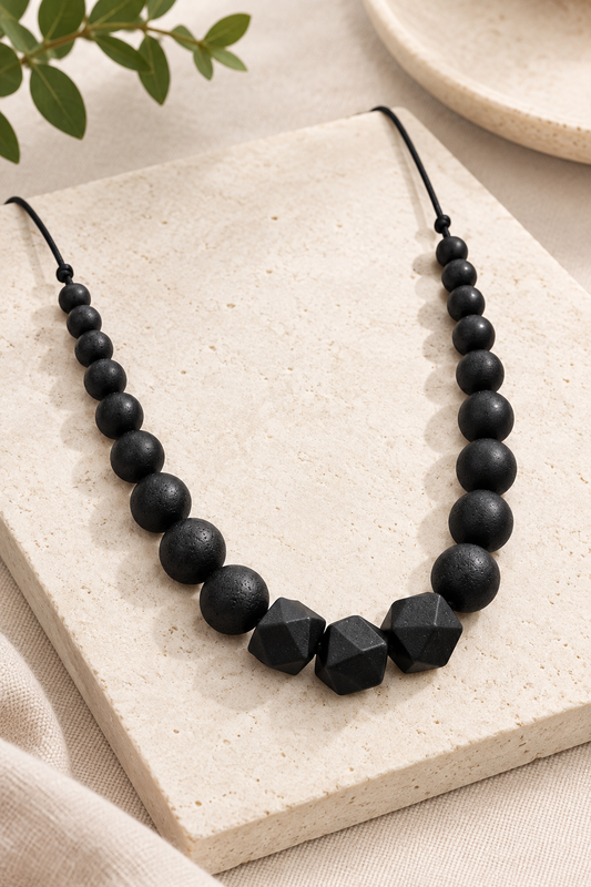 Collier d'allaitement et de portage Black Pearl : perles noires rondes et hexagonales en silicone sur cordon satin, posé sur une plaque de pierre claire